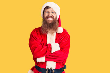 Handsome young red head man with long beard wearing santa claus costume happy face smiling with crossed arms looking at the camera. positive person.