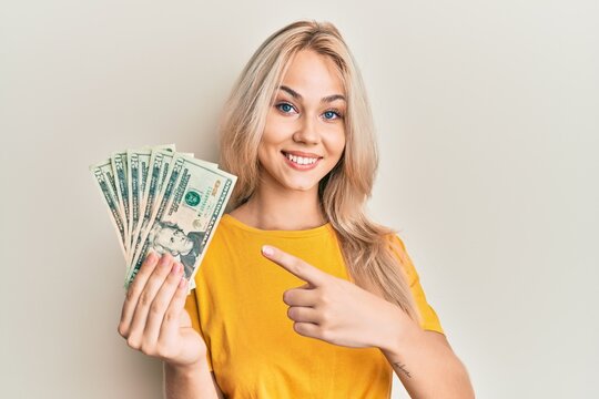 Beautiful caucasian blonde girl holding 20 dollars banknotes smiling happy pointing with hand and finger