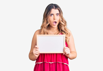 Young caucasian woman holding empty white chalkboard scared and amazed with open mouth for surprise, disbelief face