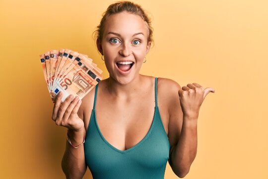 Beautiful caucasian woman holding bunch of 50 euro banknotes pointing thumb up to the side smiling happy with open mouth