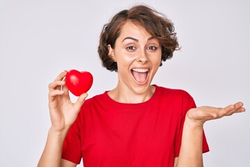 Young hispanic woman holding heart celebrating achievement with happy smile and winner expression with raised hand