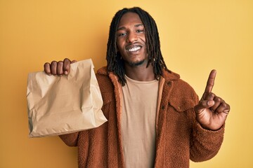 African american man with braids holding take away paper bag smiling with an idea or question pointing finger with happy face, number one