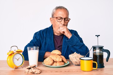 Senior handsome man with gray hair sitting on the table eating breakfast in the morning feeling unwell and coughing as symptom for cold or bronchitis. health care concept.