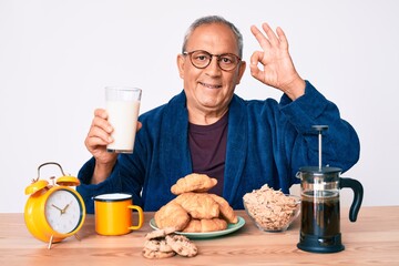 Senior handsome man with gray hair sitting on the table drinking a glass milk for breakfast doing ok sign with fingers, smiling friendly gesturing excellent symbol