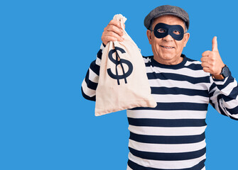 Senior handsome man wearing burglar mask holding money bag smiling happy and positive, thumb up...