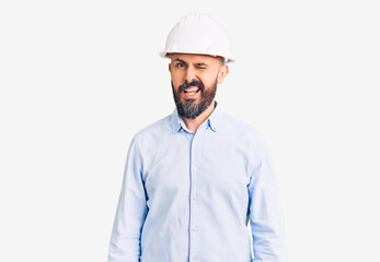 Young handsome man wearing architect hardhat winking looking at the camera with sexy expression,...