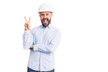 Young handsome man wearing architect hardhat smiling with happy face winking at the camera doing victory sign. number two.