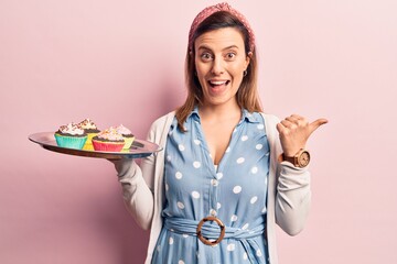 Young beautiful woman holding cupcake pointing thumb up to the side smiling happy with open mouth