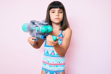 Young little girl with bang wearing swimsuit and watergun relaxed with serious expression on face....