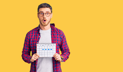 Handsome young man with bear holding travel calendar scared and amazed with open mouth for surprise, disbelief face