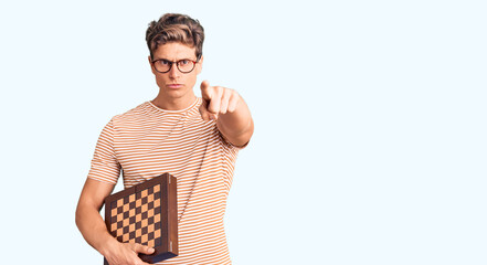 Young handsome man holding chess pointing with finger to the camera and to you, confident gesture looking serious