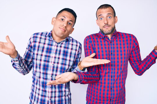 Young gay couple of two men wearing casual clothes clueless and confused expression with arms and hands raised. doubt concept.
