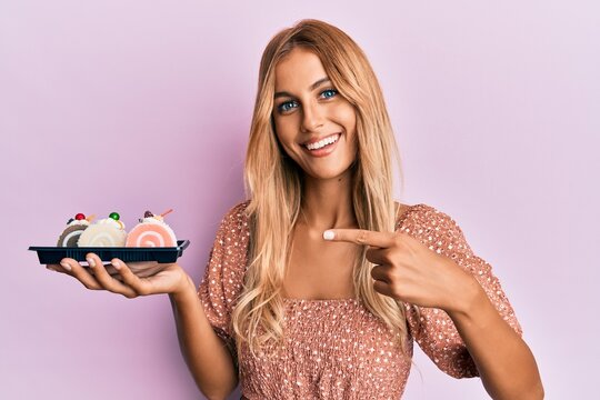 Beautiful blonde young woman holding cake sweets smiling happy pointing with hand and finger