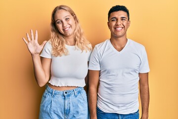 Young interracial couple wearing casual white tshirt showing and pointing up with fingers number five while smiling confident and happy.