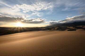 Fototapeta na wymiar Sunburst And Cloudy Mountains Over Sand Dunes