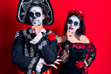 Young couple wearing mexican day of the dead costume over red looking confident at the camera with smile with crossed arms and hand raised on chin. thinking positive.