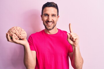 Young handsome man asking for care memory holding brain over isolated pink background smiling with...