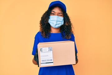 Young african american woman wearing medical mask holding delivery package winking looking at the...