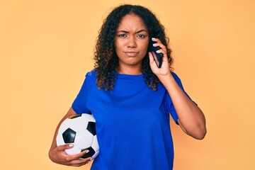 Young african american woman holding football ball talking on the smartphone skeptic and nervous, frowning upset because of problem. negative person.