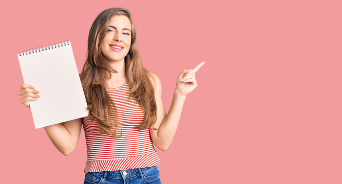 Beautiful caucasian young woman holding notebook smiling happy pointing with hand and finger to the side