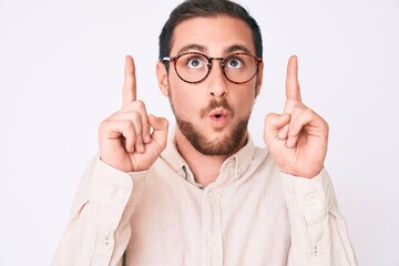 Young handsome man wearing casual clothes and glasses amazed and surprised looking up and pointing with fingers and raised arms.