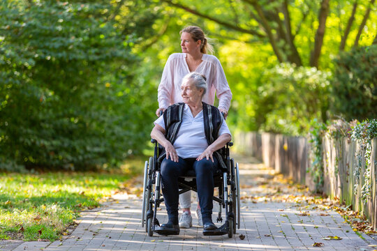 a young woman pushes an old lady in a wheelchair through a park