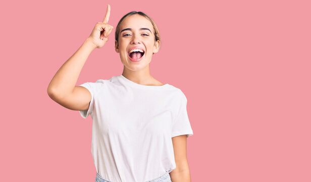 Young beautiful blonde woman wearing casual white tshirt pointing finger up with successful idea. exited and happy. number one.