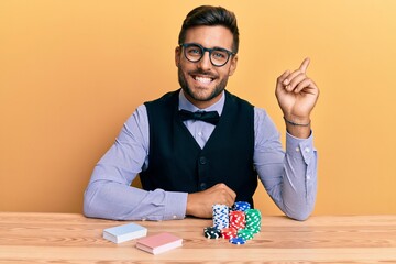 Handsome hispanic croupier man sitting on the table with poker chips and cards smiling happy...