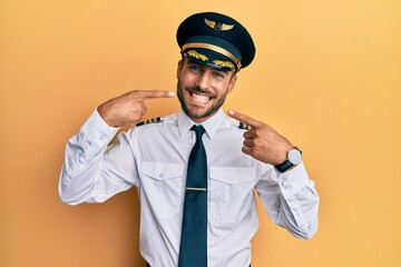 Handsome hispanic man wearing airplane pilot uniform smiling cheerful showing and pointing with fingers teeth and mouth. dental health concept.