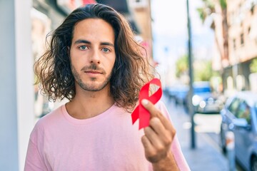 Young hispanic man with serious expression holding hiv awareness red ribbon at city. - obrazy, fototapety, plakaty