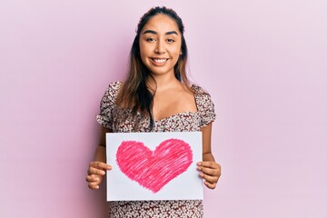 Young latin woman holding heart draw smiling with a happy and cool smile on face. showing teeth.