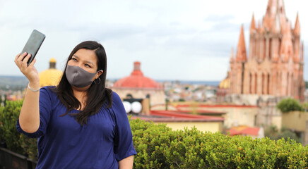 
Selfie with mobile phone of Latina tourist woman with protection mask on terrace with cathedral view in San Miguel de Allende, Guanajuato Mexico