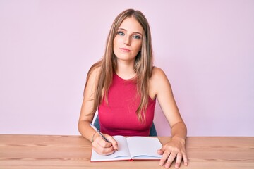 Young caucasian girl writing notebook sitting on the table thinking attitude and sober expression looking self confident