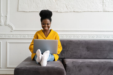 Smiling African American millennial woman with afro hairstyle wear yellow cardigan sitting on sofa, resting, looking at camera webcam and talking on a video call or skype with friends, watching movie - obrazy, fototapety, plakaty