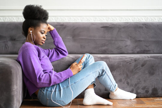 African American millennial young woman in purple sweater sitting on the floor, using smartphone, typing message, listens to music, scrolls through the feed of social media