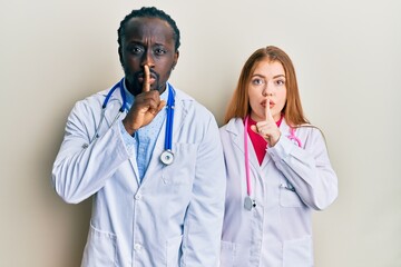 Young interracial couple wearing doctor uniform and stethoscope asking to be quiet with finger on lips. silence and secret concept.