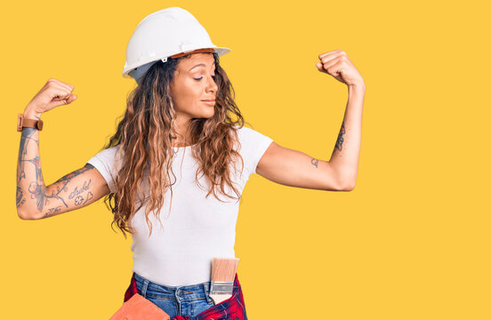 Young hispanic woman with tattoo wearing hardhat and builder clothes showing arms muscles smiling proud. fitness concept.