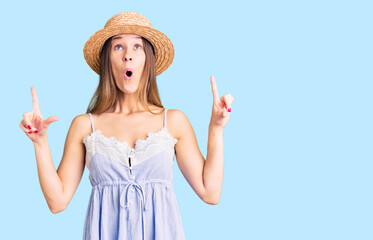 Beautiful brunette young woman wearing summer hat amazed and surprised looking up and pointing with fingers and raised arms.