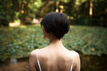 Outdoor rear view of attractive young brunette girl with short haircut admiring beautiful wild nature, spending weekend outside in park, standing in front of pond with water lilies in background