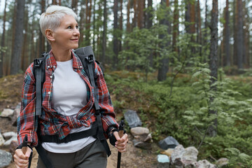 Outddor portrait of happy European female pensioner with backpack and poles, enjoying beautiful nature while nordic walking in pine forest. Aging, people, active lifestyle and health concept