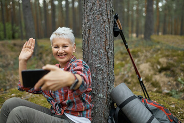 Joyful active retired female sitting under tree with hiking gear holding cell phone, smiling and waving hand, talking to her friend via video conference call using online app. People, age and travel