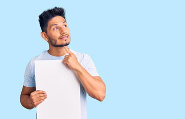 Handsome latin american young man holding cardboard banner with blank space serious face thinking...