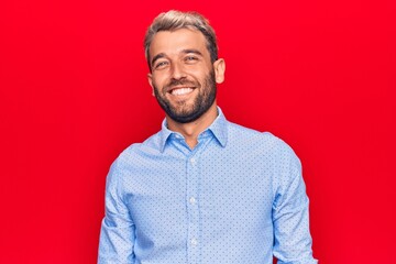 Young handsome blond man wearing casual shirt standing over isolated red background with a happy and cool smile on face. Lucky person.