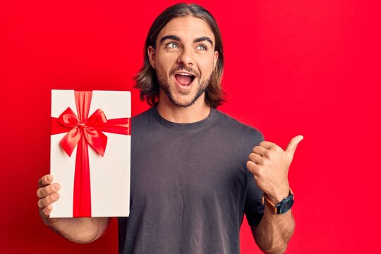 Young handsome man holding gift pointing thumb up to the side smiling happy with open mouth