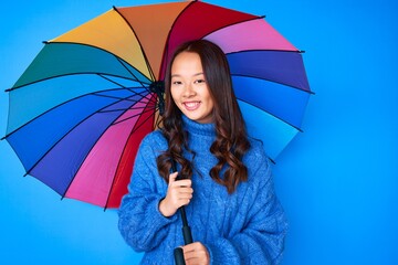 Young beautiful chinese girl holding colorful umbrella looking positive and happy standing and smiling with a confident smile showing teeth