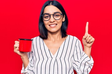 Young beautiful latin woman wearing glasses holding coffee smiling with an idea or question pointing finger with happy face, number one