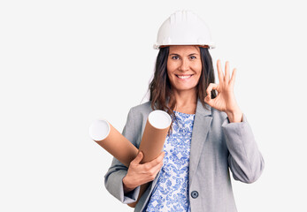 Young beautiful brunette woman wearing architect hardhat holding blueprint doing ok sign with fingers, smiling friendly gesturing excellent symbol