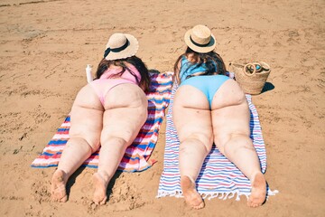 Backview of two plus size overweight sisters twins women relaxing lying on a towel at the beach on summer holidays - obrazy, fototapety, plakaty