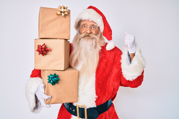 Old senior man with grey hair and long beard wearing santa claus costume holding presents screaming proud, celebrating victory and success very excited with raised arms