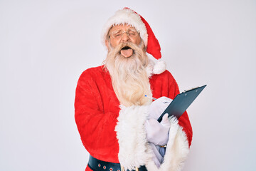 Old senior man with grey hair and long beard wearing santa claus costume writting list sticking tongue out happy with funny expression.